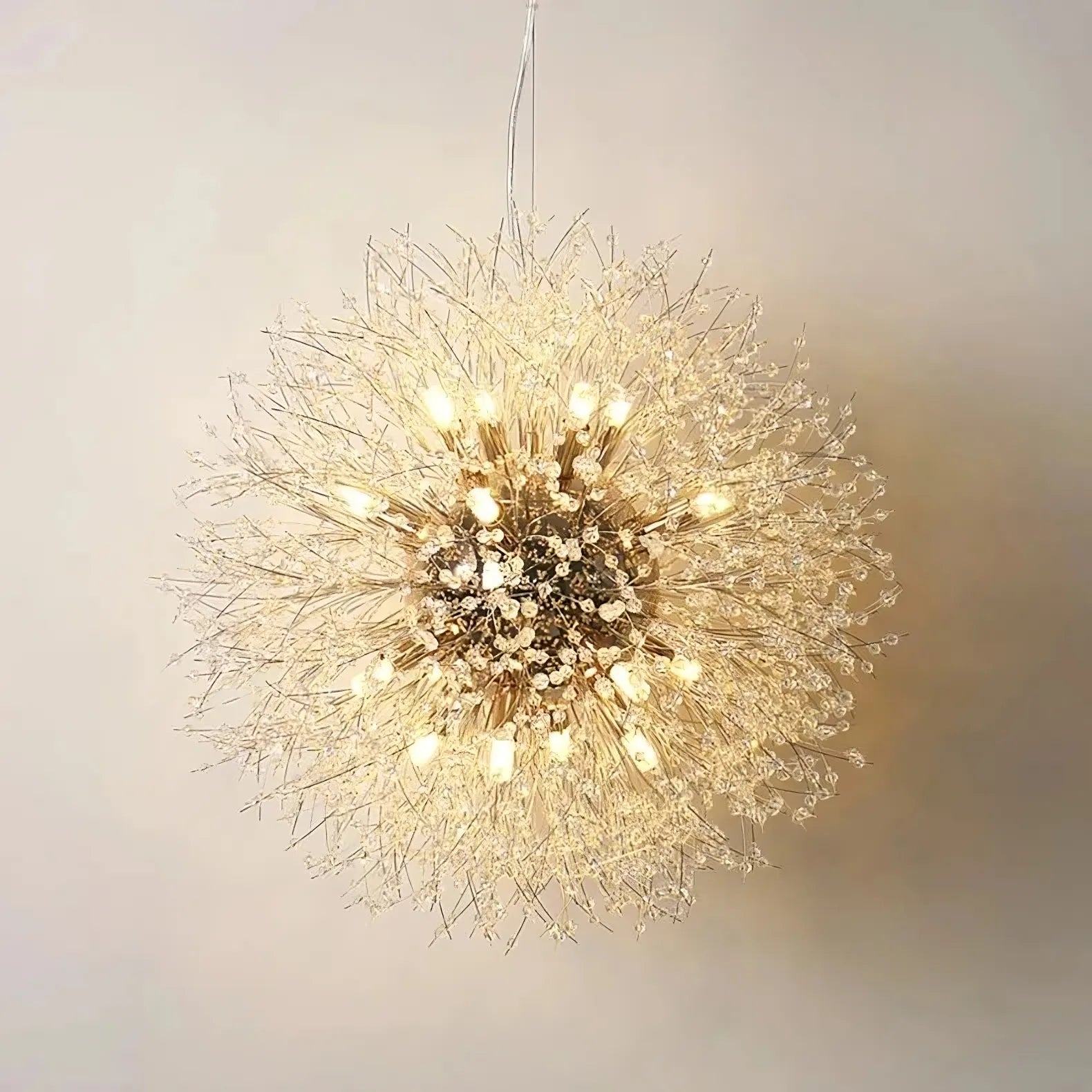 The El Sol Crystal Dandelion Lighting Fixture from Morsale.com hangs elegantly from the ceiling, resembling a delicate dandelion puff. Its numerous small and intricate branches are adorned with K9 crystals and evenly distributed light bulbs, all emitting a warm, glowing light. The stainless steel structure stands out beautifully against the plain off-white wall.