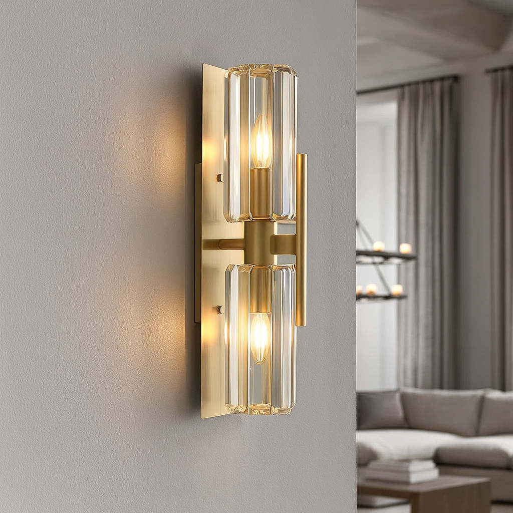 The Gio Crystal Sconce by Morsale features a modern double wall design with a gold finish and two vertical glass shades, each with a lit bulb, mounted on a light gray wall in a contemporary living room with neutral decor.