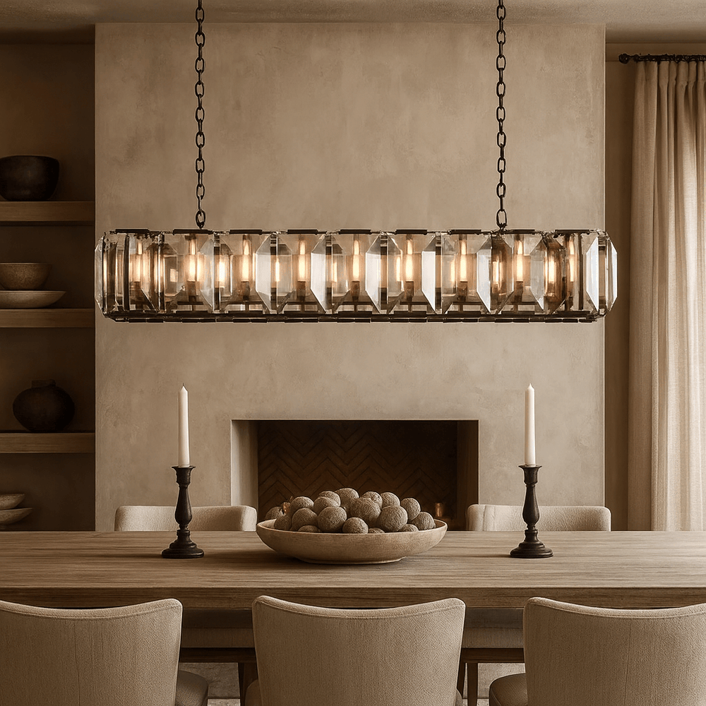 A modern dining room showcases the Brookhill Crystal Rectangular Chandelier by Morsale.com, a wooden table for eight, two candlesticks, decorative spheres in a bowl, beige walls, and a fireplace with built-in shelves.