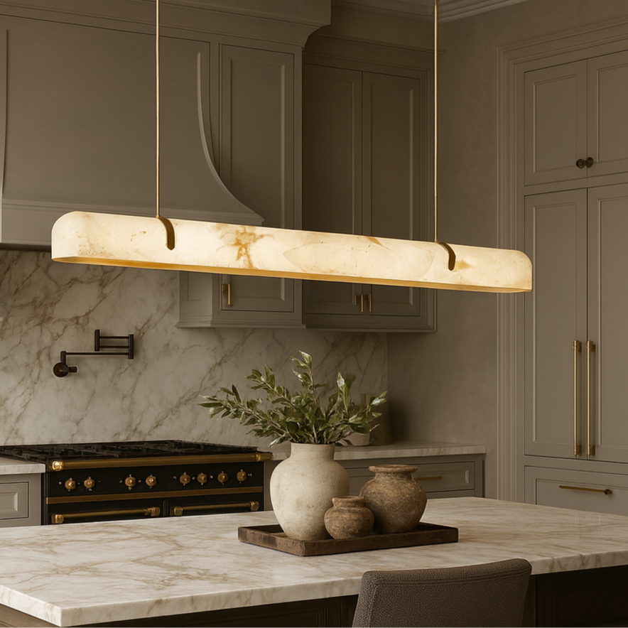 Modern kitchen with beige cabinets, marble backsplash and countertop, black and gold stove, and a Morsale Windsor Marble & Copper Linear Pendant over the island decorated with pottery and greenery.