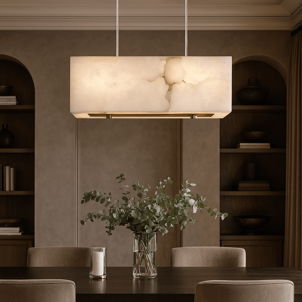 A modern dining room features the Shopp578 Leroy Marble Chandelier, a rectangular natural marble lighting pendant, above a dark wood table with beige chairs, a glass vase of eucalyptus branches, and built-in shelves in the background.