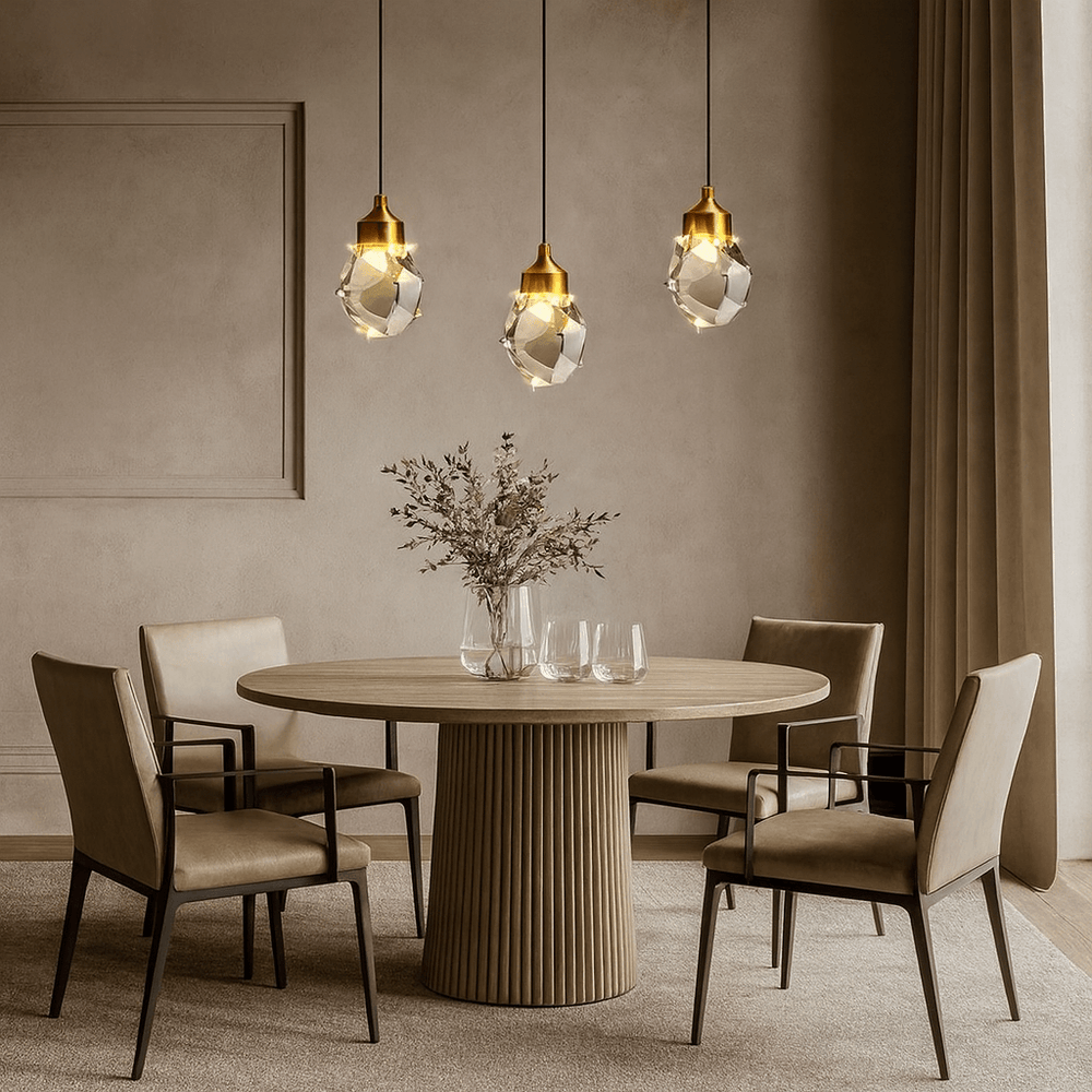 A modern dining room features a round wooden table, four beige upholstered chairs, a vase with dried flowers, and the Morsale Valenza Grand Crystal Pendant lighting hanging above beige walls with floor-length curtains.