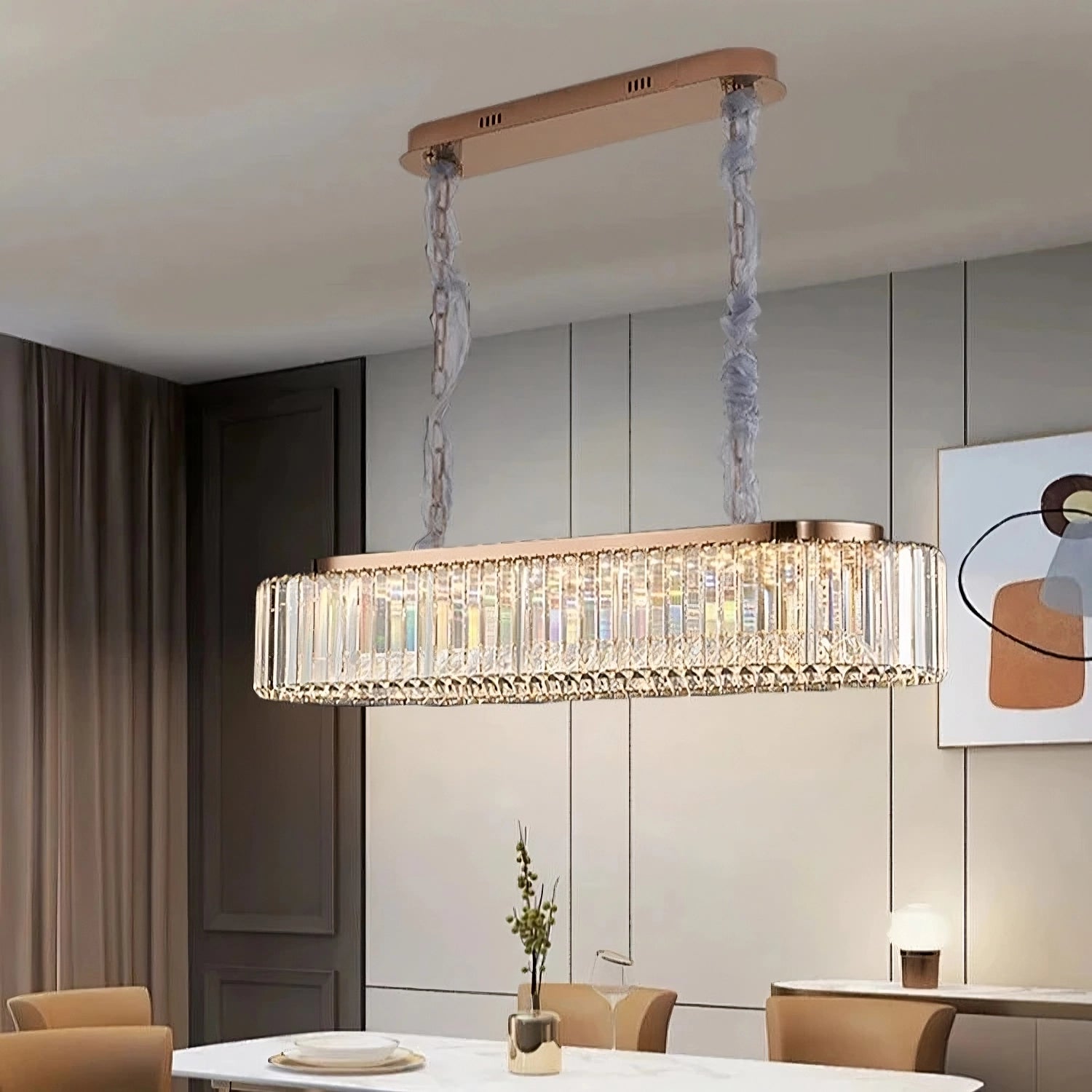 In a modern dining room, a long white table is encircled by six brown chairs. Overhead, the Gio Crystal Dining Room Chandelier by Morsale adds an elegant touch with its rectangular design and crystal accents. The beige walls display a framed abstract art piece, while natural light streams through sheer curtains.