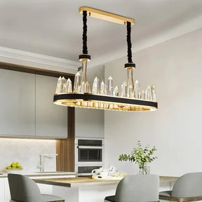 A modern kitchen features the Villa Collection Crystal Dining Room Chandelier by Morsale.com, showcasing sleek black and gold design with large hand-cut crystals hanging over a central island. The kitchen is equipped with built-in stainless steel appliances, and the light grey walls complement its chic and minimalist design.