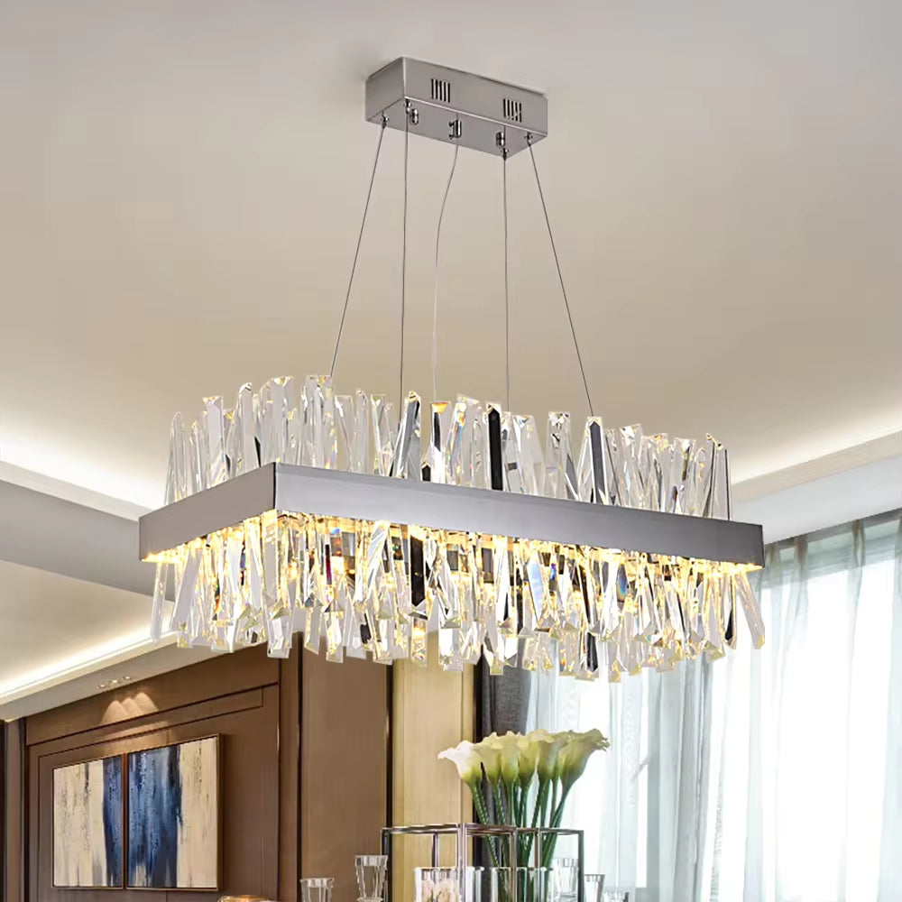 The Calvi Crystal Dining Room Chandelier by Morsale.com, a modern rectangular fixture adorned with handmade crystal accents, hangs elegantly from the ceiling on four wires. Positioned over a dining table that features a vase of white calla lilies and framed by large windows with sheer curtains, it allows natural light to flood in—creating the perfect dining room atmosphere.