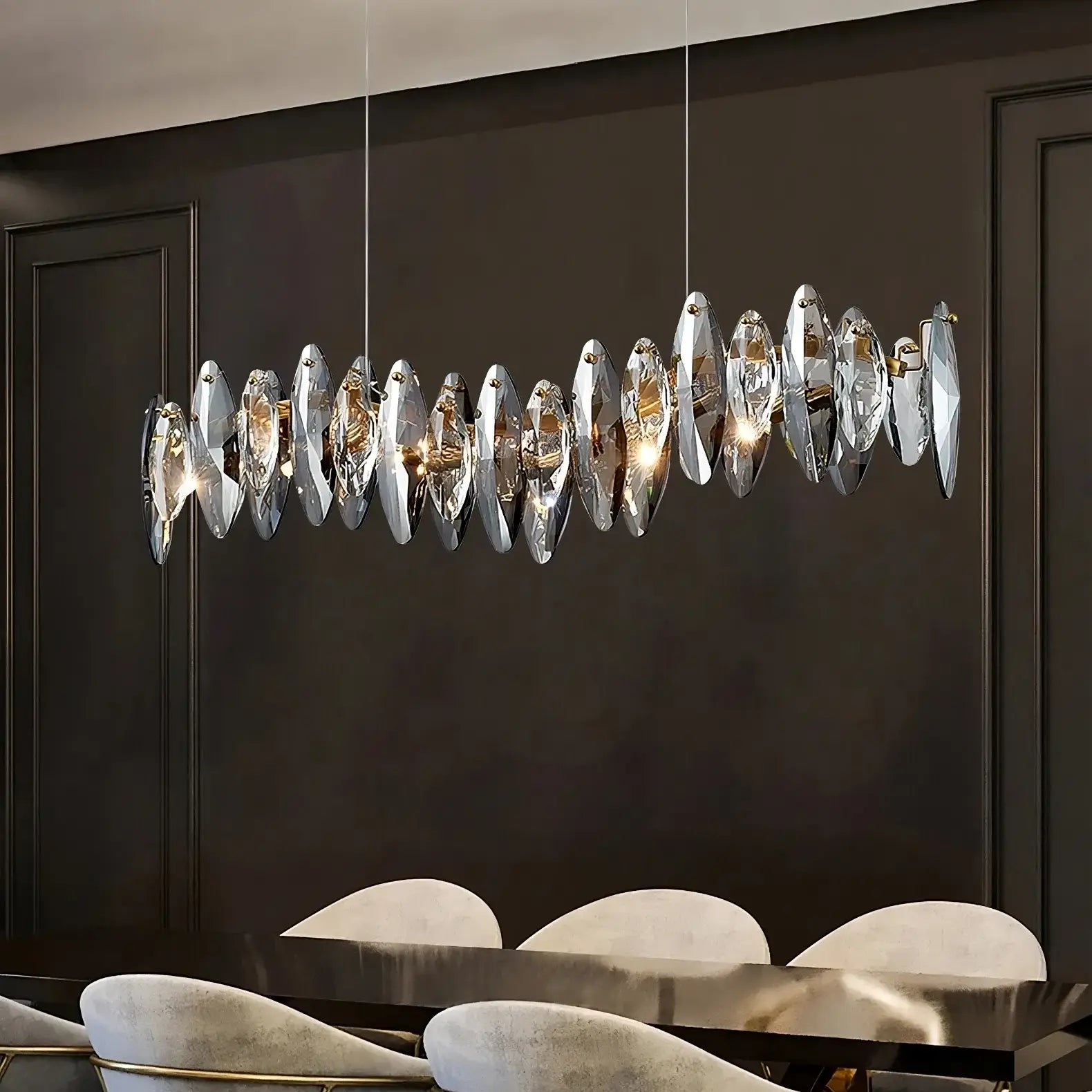 A modern dining room features the sleek, elongated Lazzo Crystal Dining Room Chandelier from Morsale.com, adorned with K9 crystal accents and equipped with energy-efficient LED bulbs, hanging over a dark wooden table. The backdrop consists of dark, sophisticated wall panels that enhance the elegant and contemporary ambiance of the space.