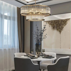 A modern dining room with a round white table set for two, featuring grey upholstered chairs. Above hangs the Bigman Gerano Modern Chandelier with LED bulbs and intricate glass detailing. A large abstract art piece adorns the wall, and a tall vase with branches serves as the table's centerpiece.