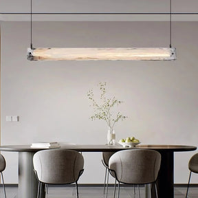 A modern luxury dining area features the Costa Dining Room Linear Suspension Chandelier by Morsale with LED bulbs illuminating a dark wooden table. Surrounded by gray cushioned chairs, the table is adorned with a vase of branches and apples. Light gray walls enhance the minimalist ambiance.