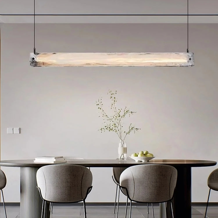 A modern luxury dining area features the Costa Dining Room Linear Suspension Chandelier by Morsale with LED bulbs illuminating a dark wooden table. Surrounded by gray cushioned chairs, the table is adorned with a vase of branches and apples. Light gray walls enhance the minimalist ambiance.