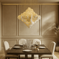A modern dining room with a dark wood table for four, cream upholstered chairs, and beige paneled walls. The Arden Marble Pendant Chandelier by Morsale.com casts warm light above, while pampas grass in a vase accents the corner.