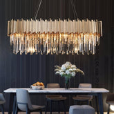 A modern dining room featuring a large, rectangular table with orchids and a fruit bowl, lit by a striking Gio Crystal Dining Room Chandelier from Morsale.com with hanging crystals against a dark wood panel wall.