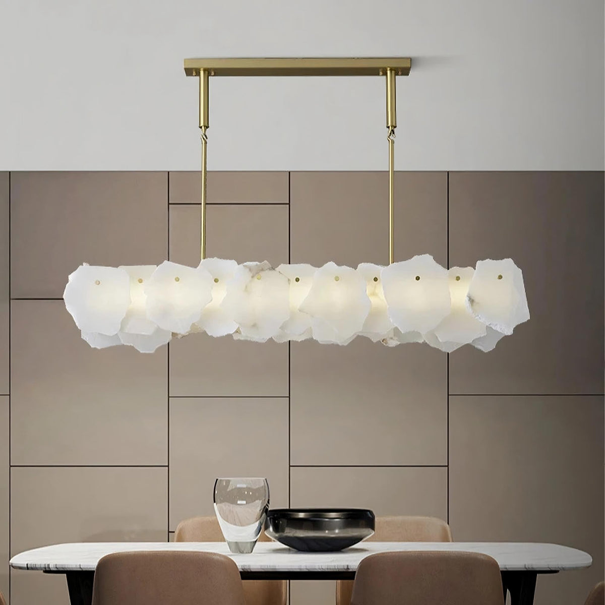 A modern dining area featuring the Natural Marble Dining Room Chandelier by Morsale, a long, rectangular fixture with a hexagonal frosted glass design hanging over a white oval table adorned with a black bowl and clear vase. The space is surrounded by beige upholstered chairs and set against a minimalist tiled wall.