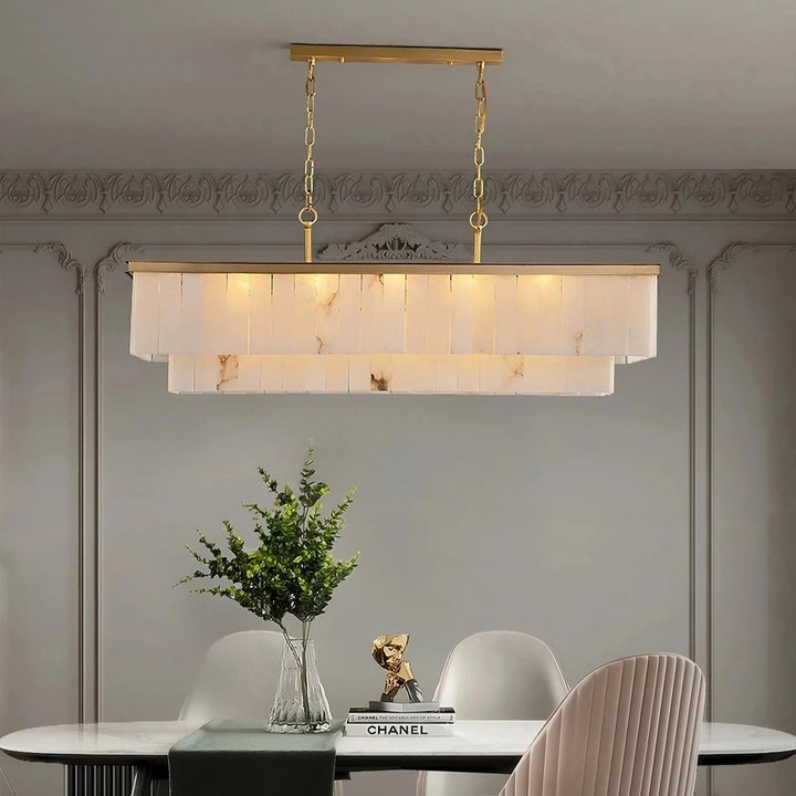 A modern dining room featuring a  Natural Calcite Crystal Dining Room Chandelier** with white and gold accents hanging above a white oval table. The table is set with a clear vase holding green foliage, next to two books and a golden decorative piece. Four chairs surround the table.