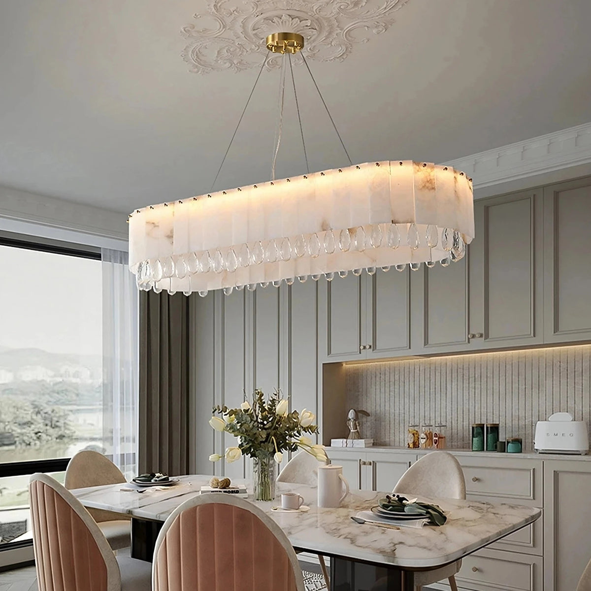 A modern dining room featuring a rectangular Spanish marble table with four pink cushioned chairs. Above the table hangs a **Morsale Natural Marble & Crystal Dining Room Chandelier**. The backdrop includes a large window with a scenic view and a built-in kitchen area with beige cabinetry.