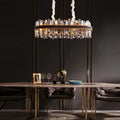 An elegant dining room is adorned with a Calvi Modern Dining Room Chandelier by Morsale.com featuring crystal and gold accents. A modern dining table with gold legs is set with wine glasses, a framed photo, a flower arrangement, and tableware. The room has dark, sophisticated decor.