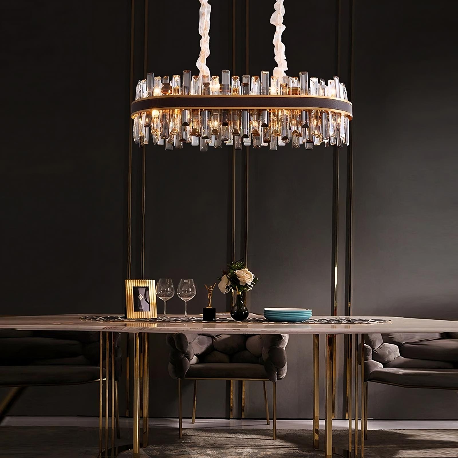 An elegant dining room is adorned with a Calvi Modern Dining Room Chandelier by Morsale.com featuring crystal and gold accents. A modern dining table with gold legs is set with wine glasses, a framed photo, a flower arrangement, and tableware. The room has dark, sophisticated decor.