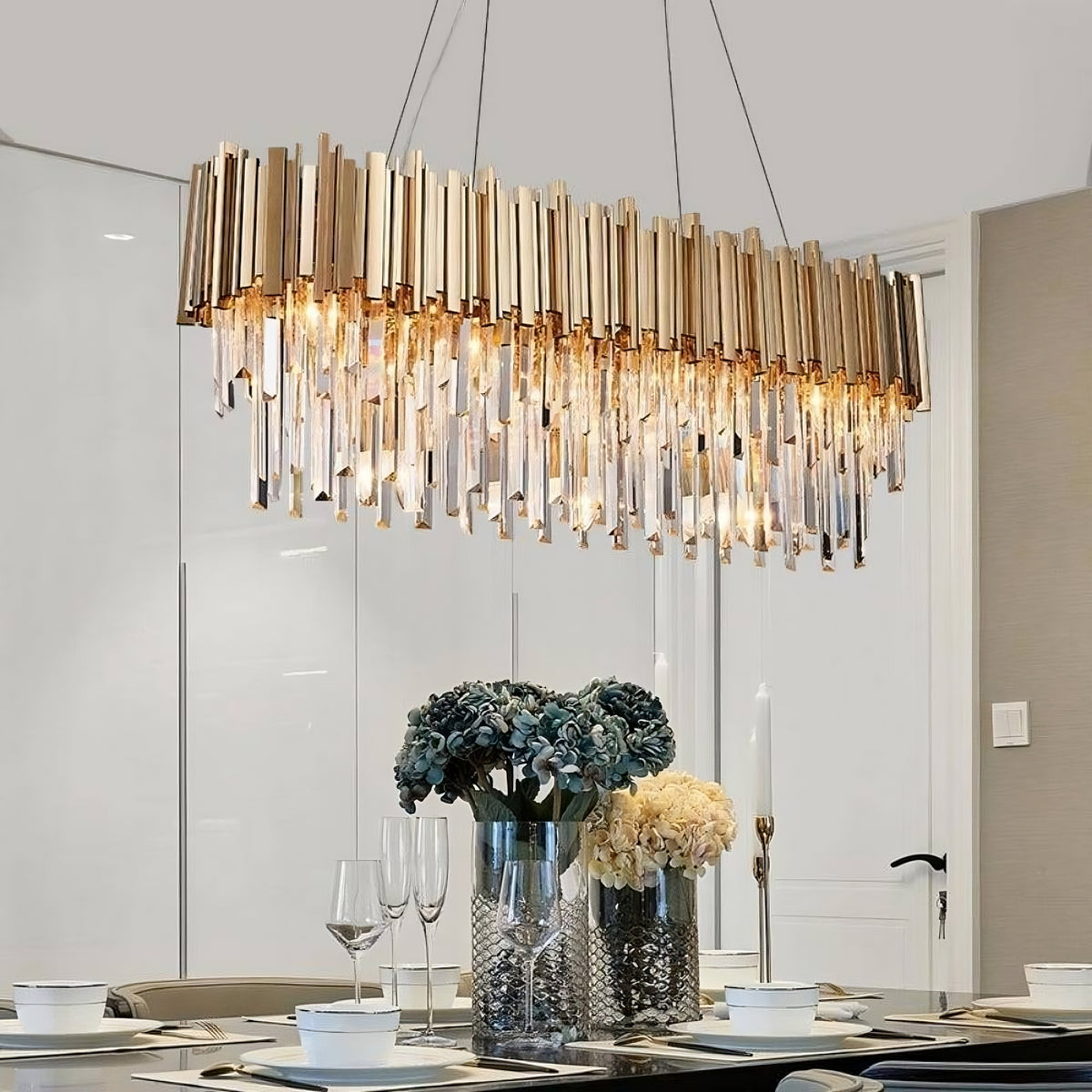 A modern dining room with a large, elegant Gio Crystal Dining Room Chandelier made of hanging gold and crystal elements over a table decorated with a vase of blue and cream flowers and empty wine glasses from Morsale.com.