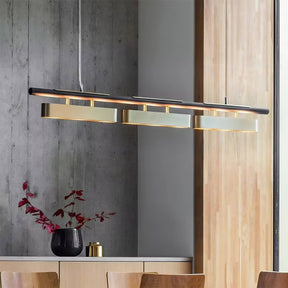 A modern dining area featuring the Marini Brass Dining Room Light Fixture from Morsale.com, a sleek hanging light fixture with three rectangular pendant lights adorned with handmade crystals. Below the light fixture, a wooden dining table holds a black vase containing branches with red leaves. The background includes wooden and concrete walls.