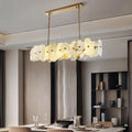 A modern dining room showcases the Morsale Norham Marble Linear Chandelier with translucent, flower-inspired glass shades above a set table, all set against neutral walls and shelves.