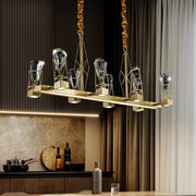 The Villa 8-Light Dining Room Light Fixture by Bigman is a modern luxury chandelier featuring a rectangular gold frame and eight geometric crystal light fixtures. The background showcases a marble backsplash, wooden cutting board, wine bottle, wine glass, metal cookware, and a closed curtain.