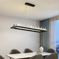 A modern dining room highlights a sleek Villa Crystal Dining Room Chandelier by Morsale.com with vertical lights above a marble table, surrounded by dark upholstered chairs. A vase with branches adorns the table, and large windows provide breathtaking outdoor views.