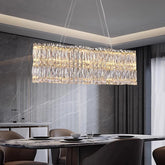 A modern dining room features the luxurious Bacci Crystal Chandelier For Dining Room by Morsale.com, suspended above a marble dining table. Neutral-toned chairs surround the table, and minimalist decor creates an elegant atmosphere. Large windows with sheer curtains allow natural light to filter in, showcasing the stunning luxury lighting.