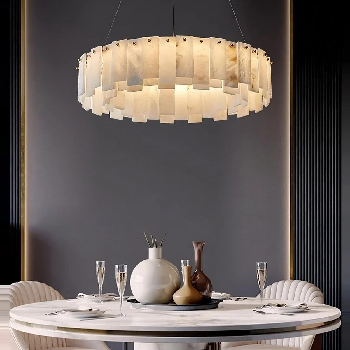 A modern dining room showcases a round white table set for four, adorned with sophisticated glassware and vases. Suspended above the table is the Moonshade Natural Marble Chandelier by Bigman, featuring layered translucent panels that cast a warm ambient glow against a dark accent wall.