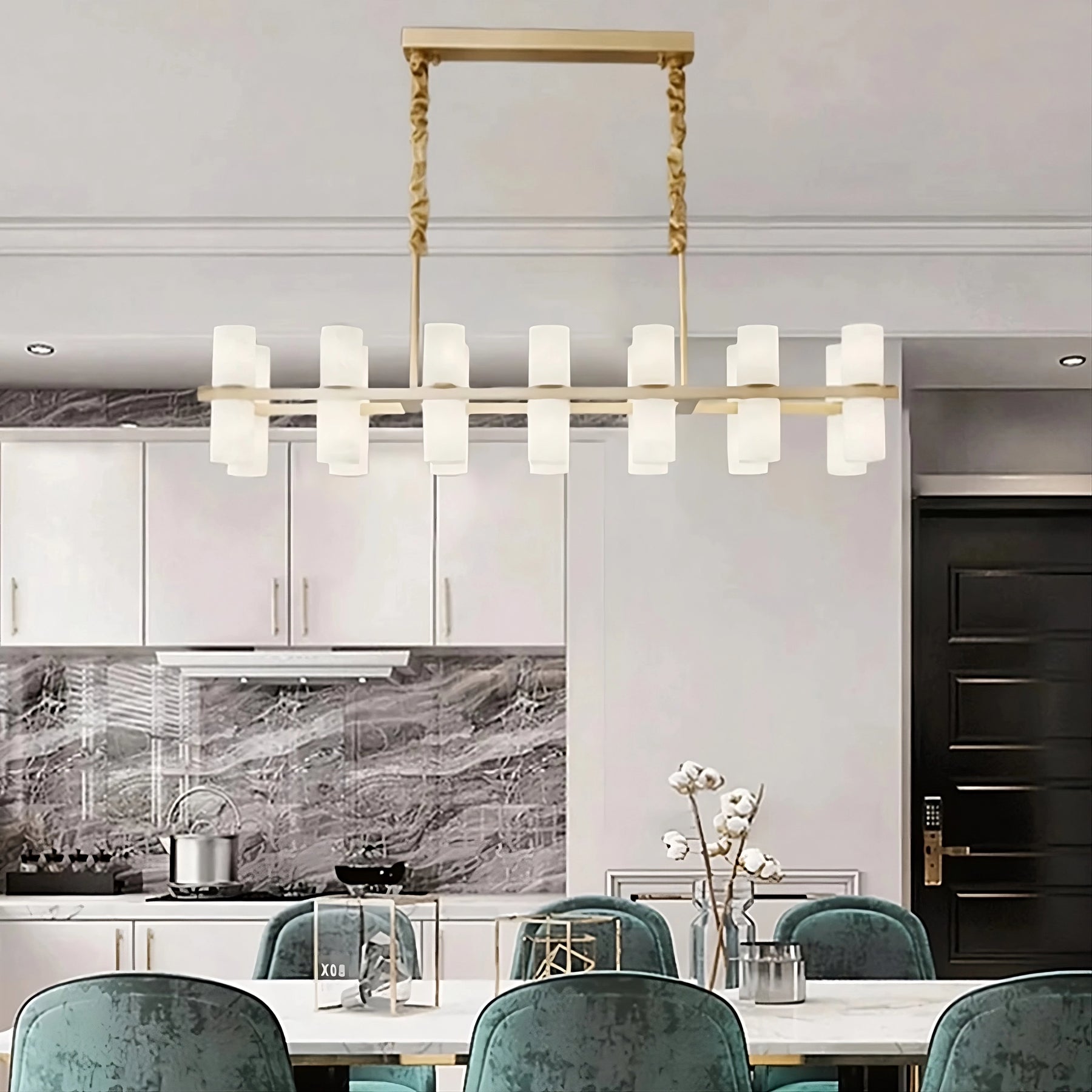 A modern dining room featuring a Morsale Moonshade Marble & Copper Chandelier casting its contemporary glow above a white table paired with six teal velvet chairs. The kitchen is outfitted with sleek polished steel appliances, white cabinets, and a marble backsplash. A vase of white flowers sits elegantly on the table.