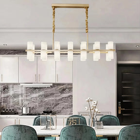 A modern dining room featuring a Morsale Moonshade Marble & Copper Chandelier casting its contemporary glow above a white table paired with six teal velvet chairs. The kitchen is outfitted with sleek polished steel appliances, white cabinets, and a marble backsplash. A vase of white flowers sits elegantly on the table.