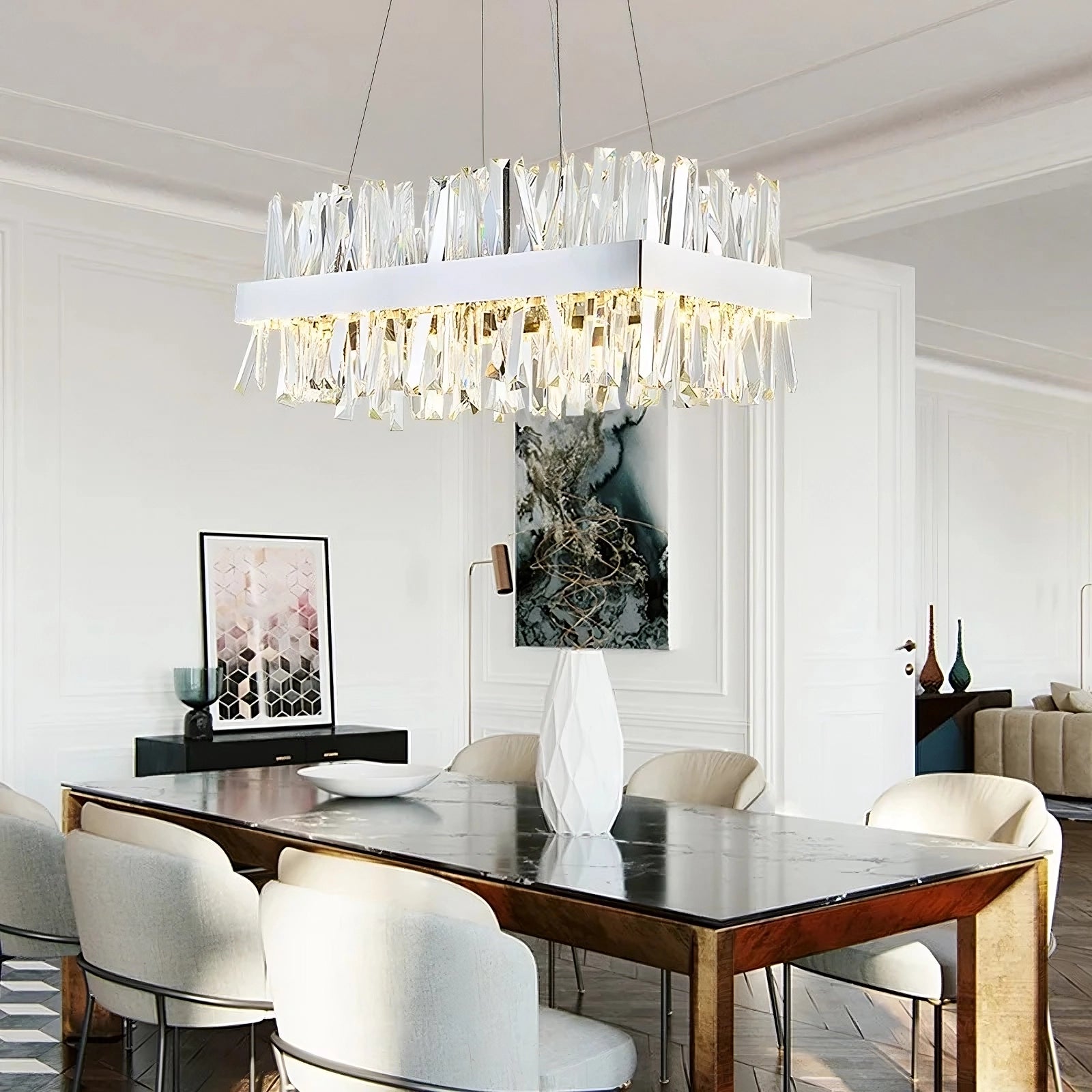 A modern dining room with a rectangular wooden table and eight white cushioned chairs. Centered above the table hangs the luxurious Calvi Crystal Dining Room Chandelier by Morsale.com, featuring handmade crystals. The room boasts white walls adorned with minimalistic decor, including a large abstract artwork and a vase on a side table.