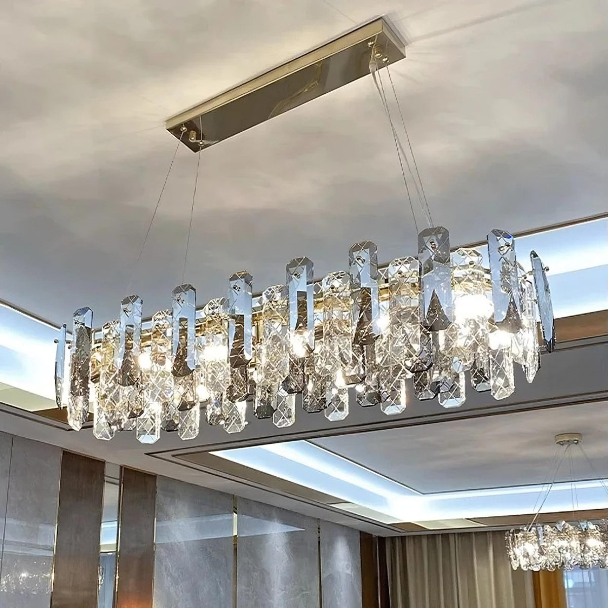 A modern rectangular Lazzo Crystal Ceiling Chandelier from Morsale.com hangs from the ceiling, featuring multiple elongated, geometric crystal pieces. This chandelier emits a bright light through energy-efficient LED lighting, creating reflections and a luxurious atmosphere in the room with a coffered ceiling and contemporary decor.