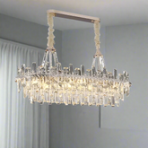 In a modern room, the Gio Crystal Chandelier for Dining Room by Morsale.com hangs from the ceiling, boasting a luxurious rectangular design. It features multiple vertical crystal rods that elegantly refract light, creating a sparkling effect. The background displays light-colored curtains and a plain wall.