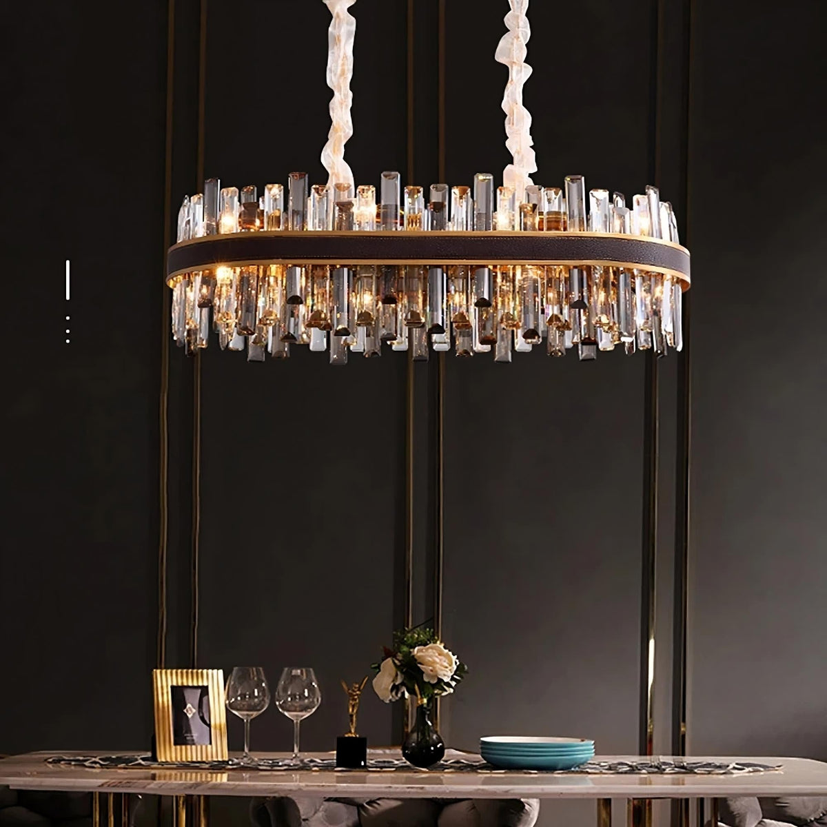 A modern dining room with a large, cylindrical Calvi Crystal Dining Room Chandelier from Morsale.com hanging from the ceiling. The chandelier features an array of clear, rectangular prisms and emits a warm glow. Below, a dining table is set with plates, glasses, a framed photo, and a flower vase.