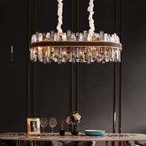 A modern dining room with a large, cylindrical Calvi Crystal Dining Room Chandelier from Morsale.com hanging from the ceiling. The chandelier features an array of clear, rectangular prisms and emits a warm glow. Below, a dining table is set with plates, glasses, a framed photo, and a flower vase.