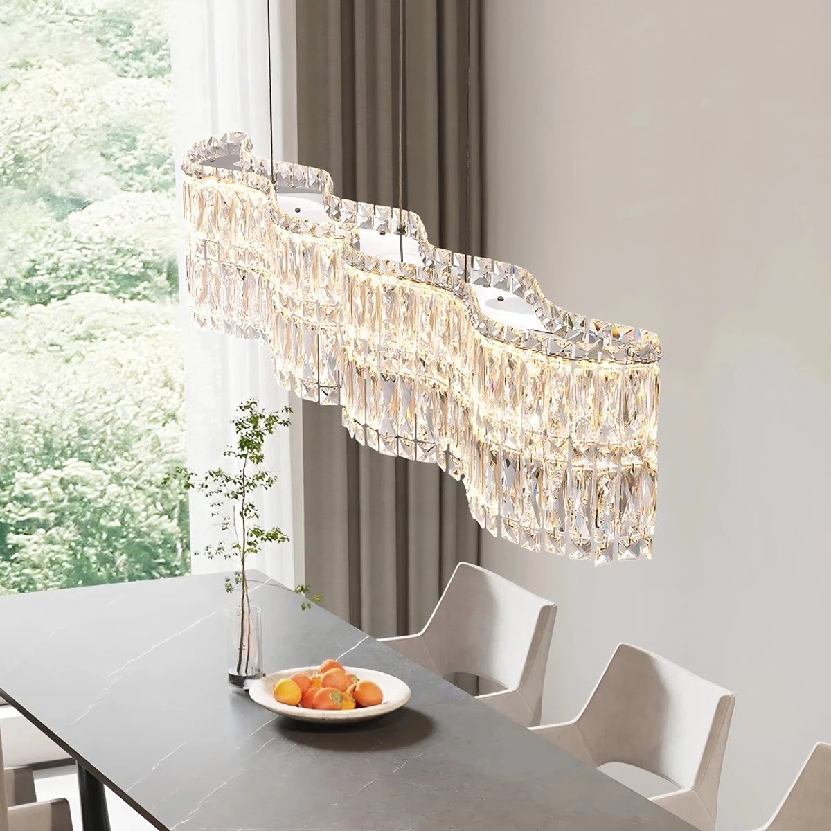 A modern dining room features the Bacci Crystal Chandelier from Morsale.com, an elegant fixture with multiple sparkling tiers, hanging over a dark gray table. On the table is a clear vase with a single leafy branch and a bowl containing various fruits. Large windows in the background reveal a lush green view, beautifully accented by luxurious lighting.