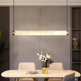 A modern dining room features the chic Moonshade Natural Marble Dining Room Light Fixture by Bigman, elegantly suspended above a round table. The table is adorned with a vase of white and brown flowers and scattered orange berries, forming an appealing contrast. Ambient lighting enhances the scene against the gray wall and shelves in the background.