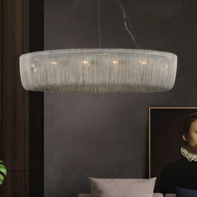 A modern Cadena Chain Tassel Chandelier Light by Morsale.com hangs from the ceiling, featuring a ribbed glass exterior. The illuminated contemporary fixture casts a warm glow in the dimly lit room. In the background, part of a framed portrait and a gray couch with cushions are visible.