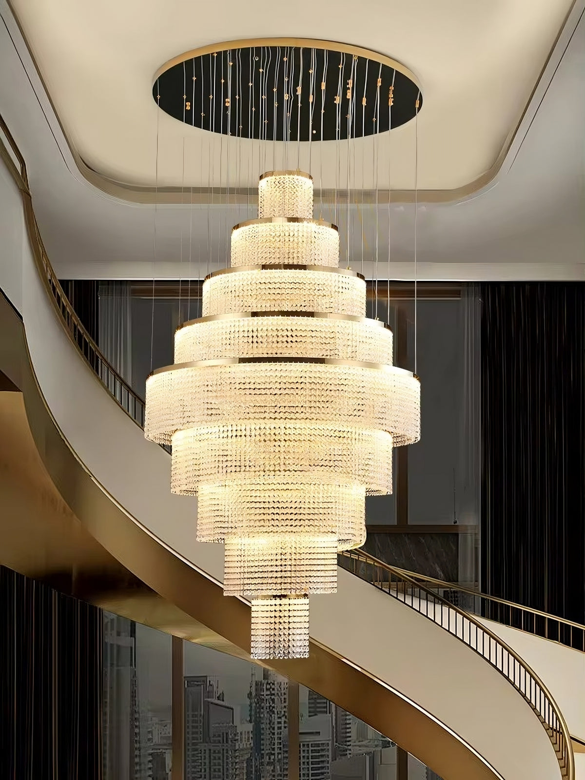 An opulent Alleri Crystal Foyer Chandelier in gold from Morsale.com graces a high ceiling with its modern circular mount. The grand multi-tiered chandelier, adorned with crystal shades and a cascading design, casts a radiant glow over the sweeping spiral staircase below. Its illumination starkly contrasts against dark walls and large windows, enhancing the luxurious interior setting and showcasing exquisite modern lighting.