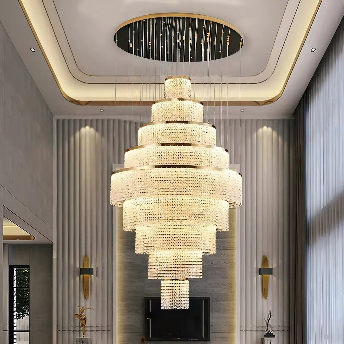 The Alleri Crystal Foyer Chandelier in Gold from Morsale.com, a luxurious and large fixture with multiple tiers of sparkling crystal shades, is suspended from a modern ceiling featuring ambient lighting. This stunning chandelier adorns a spacious, elegantly decorated room with high ceilings, large windows, and wall-mounted lights.