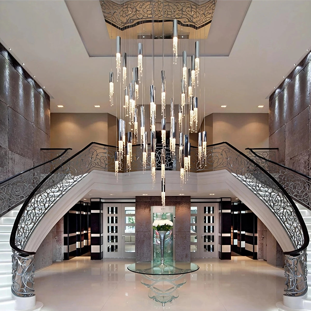 A grand, modern foyer with a double staircase and intricate railings on either side. At the center, a large, elegant Canali Modern Crystal Pendant Chandelier from Morsale.com adorned with clear crystals hangs from the ceiling. The polished floor reflects the light, and a glass table with a floral arrangement stands beneath the chandelier.