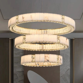 A luxurious Marble & Crystal Modern Chandelier by Morsale.com with three circular tiers, each adorned with natural marble-patterned panels and accented by crystal details, hangs from the ceiling. The background features a modern design with neutral tones and geometric decor elements.