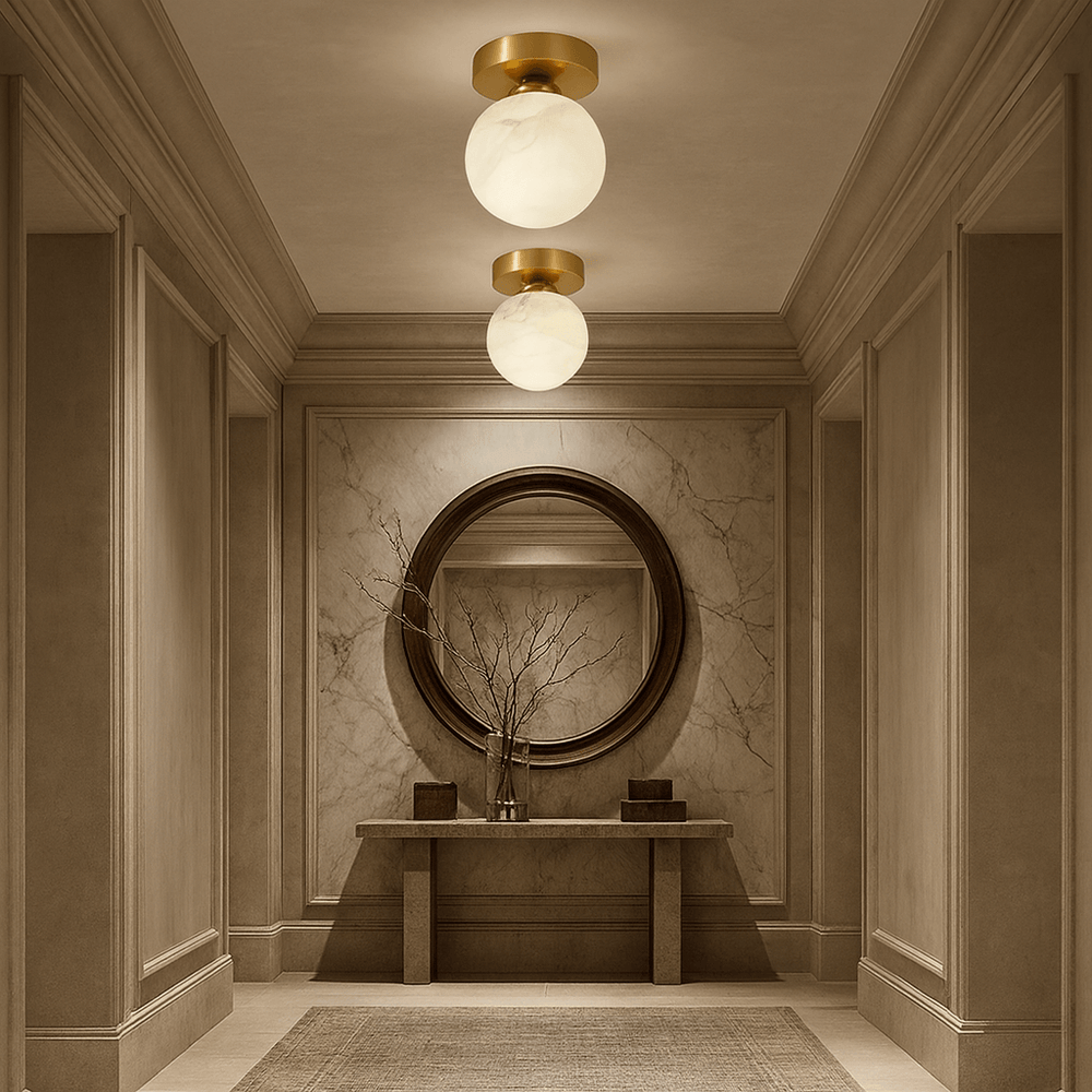 A sophisticated, warmly lit hallway features a large round mirror above a marble console, decorated with a branch-filled vase and small boxes. The Soria Marble Flushmount by Morsale.com bathes the neutral space in elegant light.