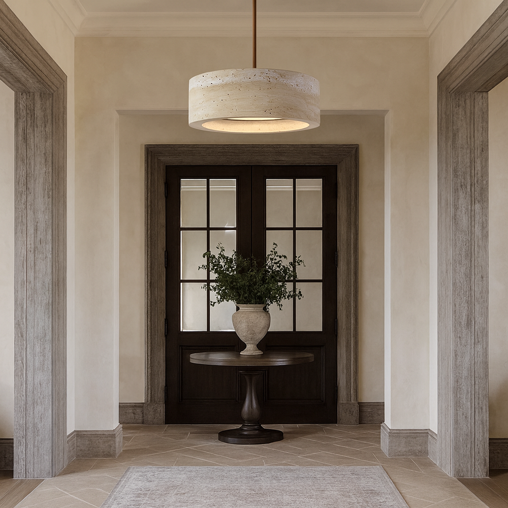 A round wooden table with a vase of greenery stands in a hallway featuring beige walls, a large dark double door with glass panes, stone tile flooring, and the Morsale Vero Travertine Bell Pendant overhead.