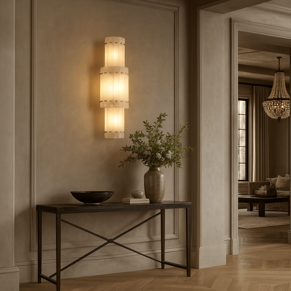 
                  
                    The Valmont Marble Wall Sconce by Morsale, softly illuminating a black console table with greenery and a bowl, is mounted on beige paneled walls in an elegant room with a chandelier in the background.
                  
                