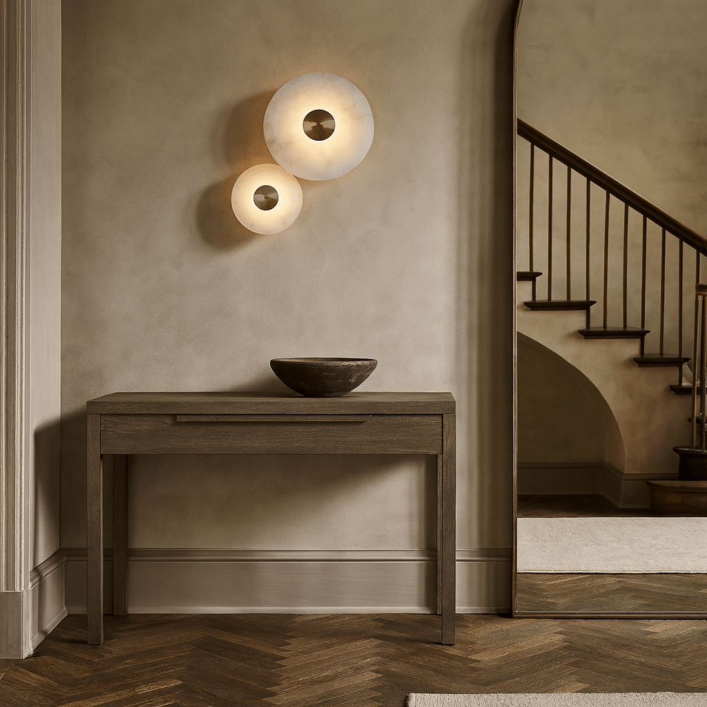A minimalist entryway features a wooden console table, a dark ceramic bowl, two Solena Marble Disc Wall Sconces from Morsale.com emitting soft light, and a staircase visible through an arched doorway in the background.