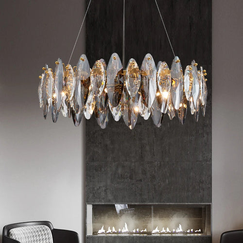 The Lazzo Modern Chandelier by Morsale.com, featuring gold and handmade leaf-shaped crystals, hangs above a contemporary fireplace against a textured dark wall, with a gray upholstered chair in the foreground.