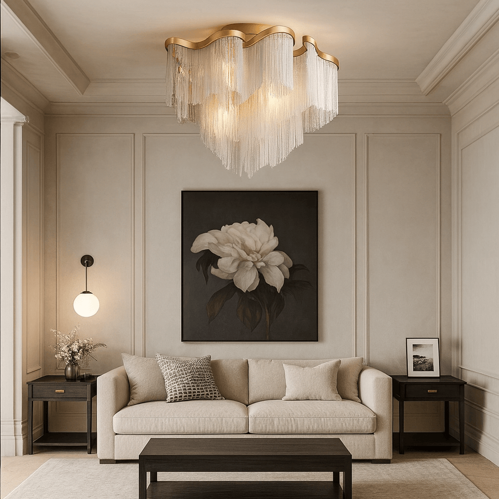 A modern living room features a beige sofa, dark wood tables, floral art, and the Cadena Chain Tassel Ceiling Light Fixture from Morsale.com, with gold cascading fringe. Light walls, a round wall lamp, and a small flower vase complete the minimal decor.