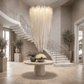 A luxurious foyer features a large round stone table with white and green flower vases, potted trees, and the Alleri Luxury Staircase Chandelier by Morsale.com cascading between two curved marble staircases.