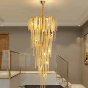 An elegant *Albero Crystal Foyer Chandelier* by Morsale.com with cascading crystal-like strands hangs in a spacious, modern room with high ceilings. The room features a glass and metal stair railing, minimalistic décor, and a blend of neutral tones, enhancing the polished gold chandelier's radiant glow.