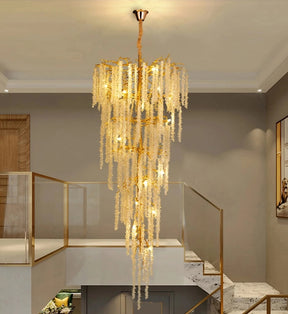 An elegant *Albero Crystal Foyer Chandelier* by Morsale.com with cascading crystal-like strands hangs in a spacious, modern room with high ceilings. The room features a glass and metal stair railing, minimalistic décor, and a blend of neutral tones, enhancing the polished gold chandelier's radiant glow.